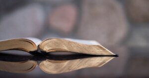 A Bible lies open, reflected by the glass tabletop.