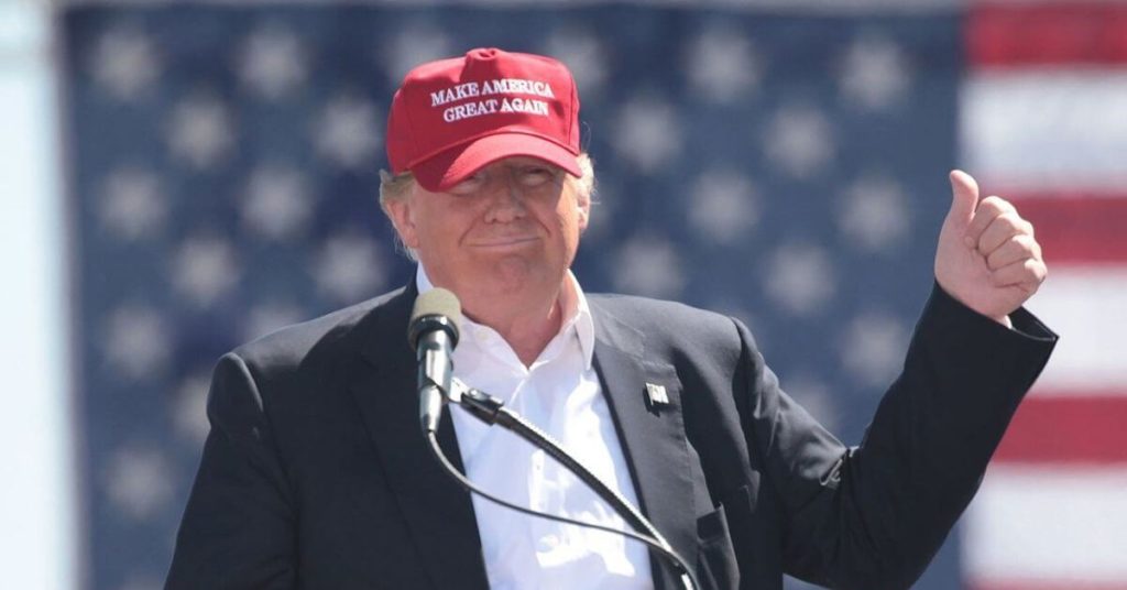 Trump standing in front of an American flag wearing a MAGA hat and giving a thumb's up