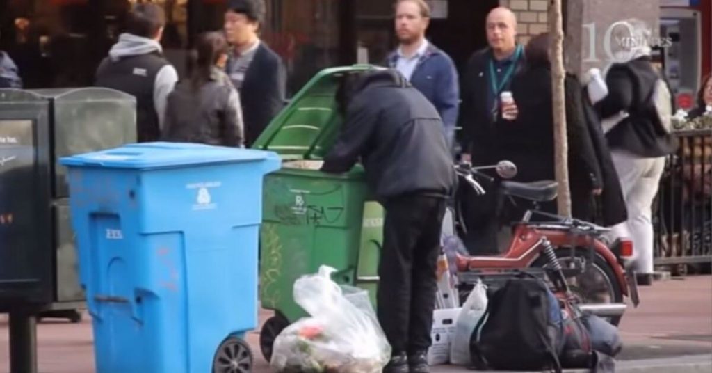 A homeless person digging in a trash can on a busy street.