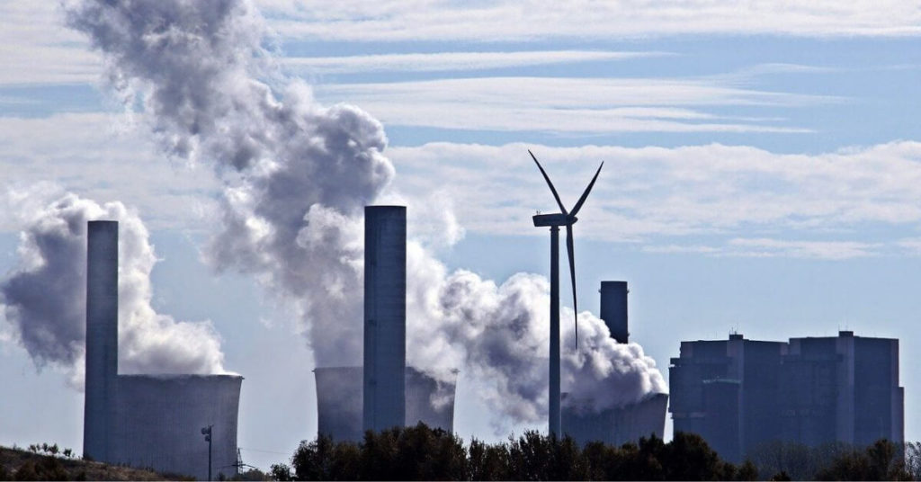 A photograph of a single wind turbine in front of a line of factory buildings billowing smoke.