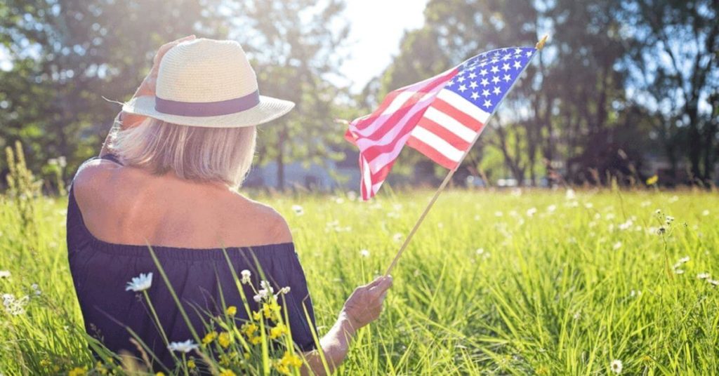 A photograph of a woman waving an American flag while sitting in a sunny field.