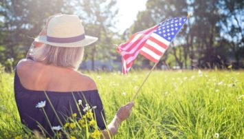 A photograph of a woman waving an American flag while sitting in a sunny field.