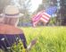 A photograph of a woman waving an American flag while sitting in a sunny field.