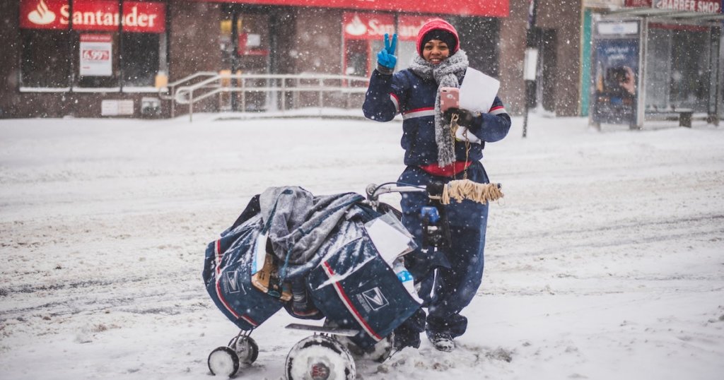 A photograph of a letter carrier in the snow with an overloaded trolley of mail. She is giving the peace sign.