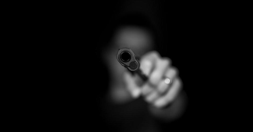 A hand points a gun at the viewer from out of the shadows.